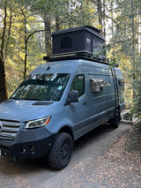 A rugged grey Mercedes Sprinter rv rental, featuring a roof-top tent, ladder, and off-road tires, parked in a sunny forest.