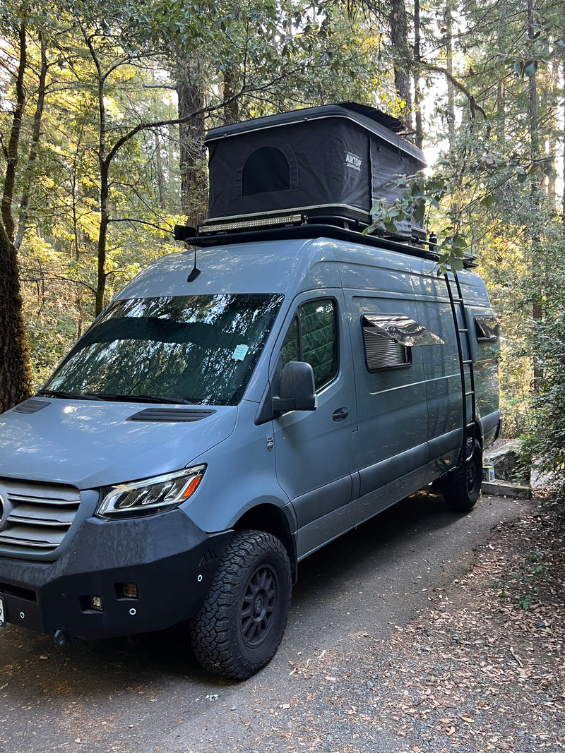 A rugged grey Mercedes Sprinter rv rental, featuring a roof-top tent, ladder, and off-road tires, parked in a sunny forest. — photo 8