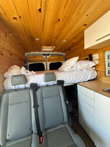 Interior of a cozy RV rental: wood-paneled walls, bed, kitchenette, and passenger seats.