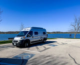 A silver camper van (RV rental) parked by a scenic blue lake under a clear sky. Perfect for a road trip adventure!