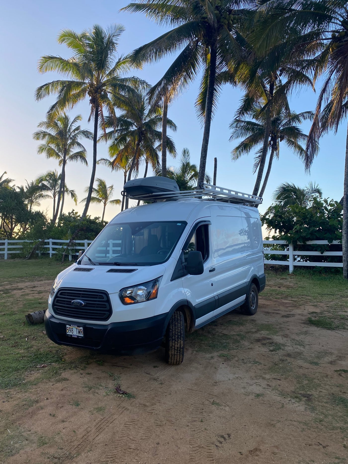 White adventure van with roof storage and rack parked on dirt amid tall palm trees and a white fence. RV rental ready! — photo 15