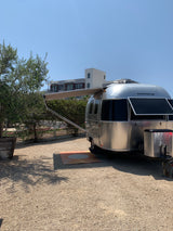 A sleek silver Airstream rv rental is parked on a sunlit gravel site with its awning extended for shade.