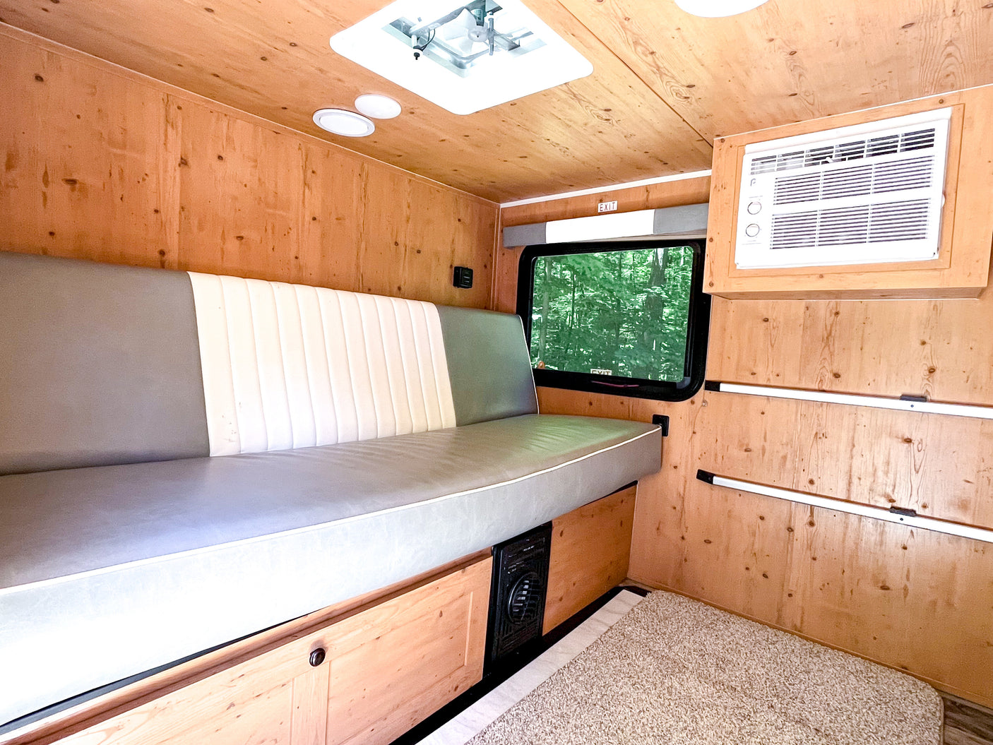 Inside a cozy **RV rental**: wood-paneled walls/ceiling, bench bed, AC unit, window with forest view, and carpeted floor.