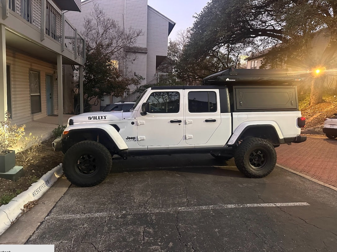 White Jeep Gladiator with black bed camper & roof-top tent, parked outside. A versatile rv rental for adventures.