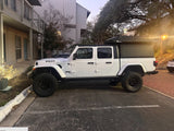 White Jeep Gladiator with black bed camper & roof-top tent, parked outside. A versatile rv rental for adventures.