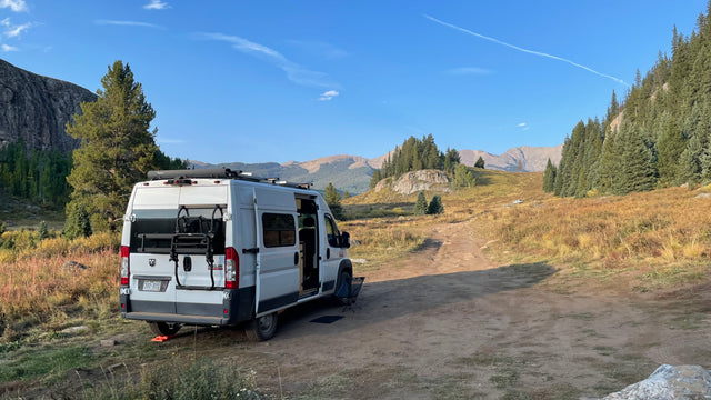 White RV rental van parked on a dirt road in a scenic mountain landscape. Trees, dry grass, and peaks under blue sky.