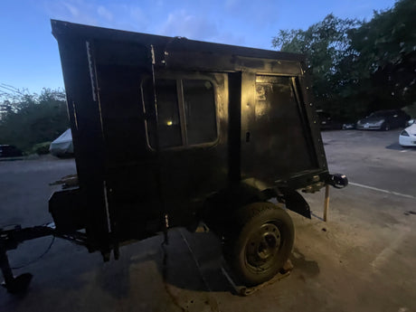 Dusk view of a rustic black custom trailer, resembling a small camper or DIY RV rental. Parked on asphalt with trees & cars.