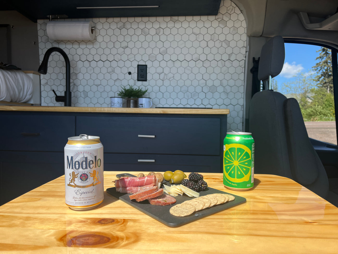 Cozy RV rental setup: a wooden table with charcuterie, Modelo beer, and lime soda, overlooking the van's kitchen.