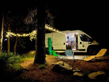Night view of an RV rental campsite: bright van interior, string lights, and outdoor chairs amidst trees, creating a cozy scene.
