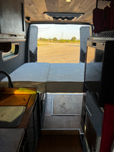 Inside a functional RV rental camper van: bed, kitchen area with sink, and open rear doors revealing a scenic outdoor view.