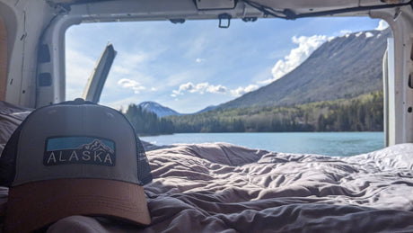 RV rental view: An "Alaska" hat on a cozy bed overlooks a clear lake & majestic mountains.
