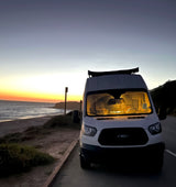 An RV rental van, a white Ford Transit, parked by a scenic beach at sunset with glowing interior lights, ready for adventure.
