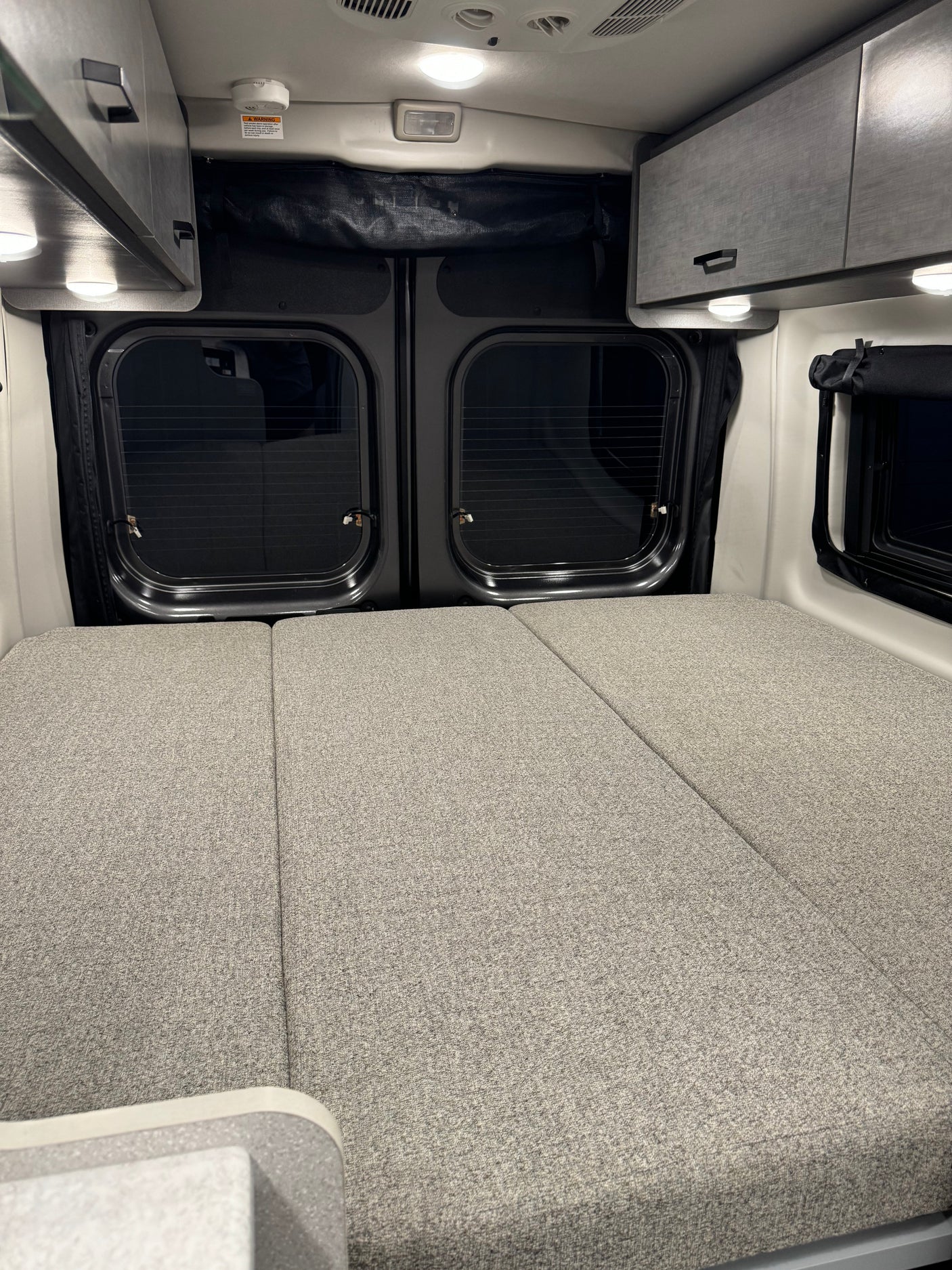 Comfortable RV rental interior featuring a spacious bed, overhead storage cabinets, and rear windows, ready for your trip.