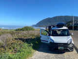 White camper van (RV rental) on the scenic Oregon coast, parked with its awning extended, overlooking the ocean & mountains.