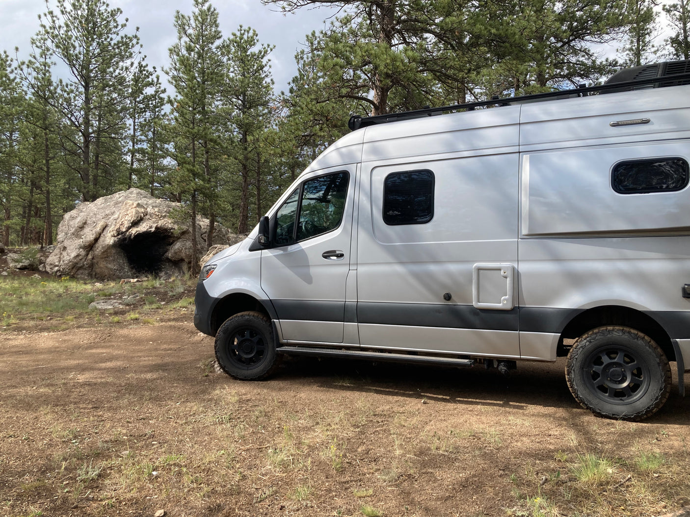 Silver RV rental camper van parked on a dirt road in a pine forest with a large boulder. Ready for off-road adventures.