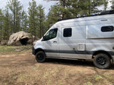 Silver RV rental camper van parked on a dirt road in a pine forest with a large boulder. Ready for off-road adventures.
