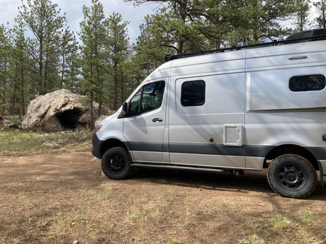 Silver RV rental camper van parked on a dirt road in a pine forest with a large boulder. Ready for off-road adventures.