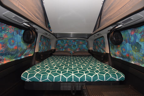 Inside an RV rental: a cozy bed with geometric blanket, ocean-themed curtains, and fans for comfort.