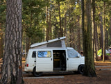 An RV rental pop-top camper van at a sunny forest campsite with its side door open, surrounded by tall trees and tents.