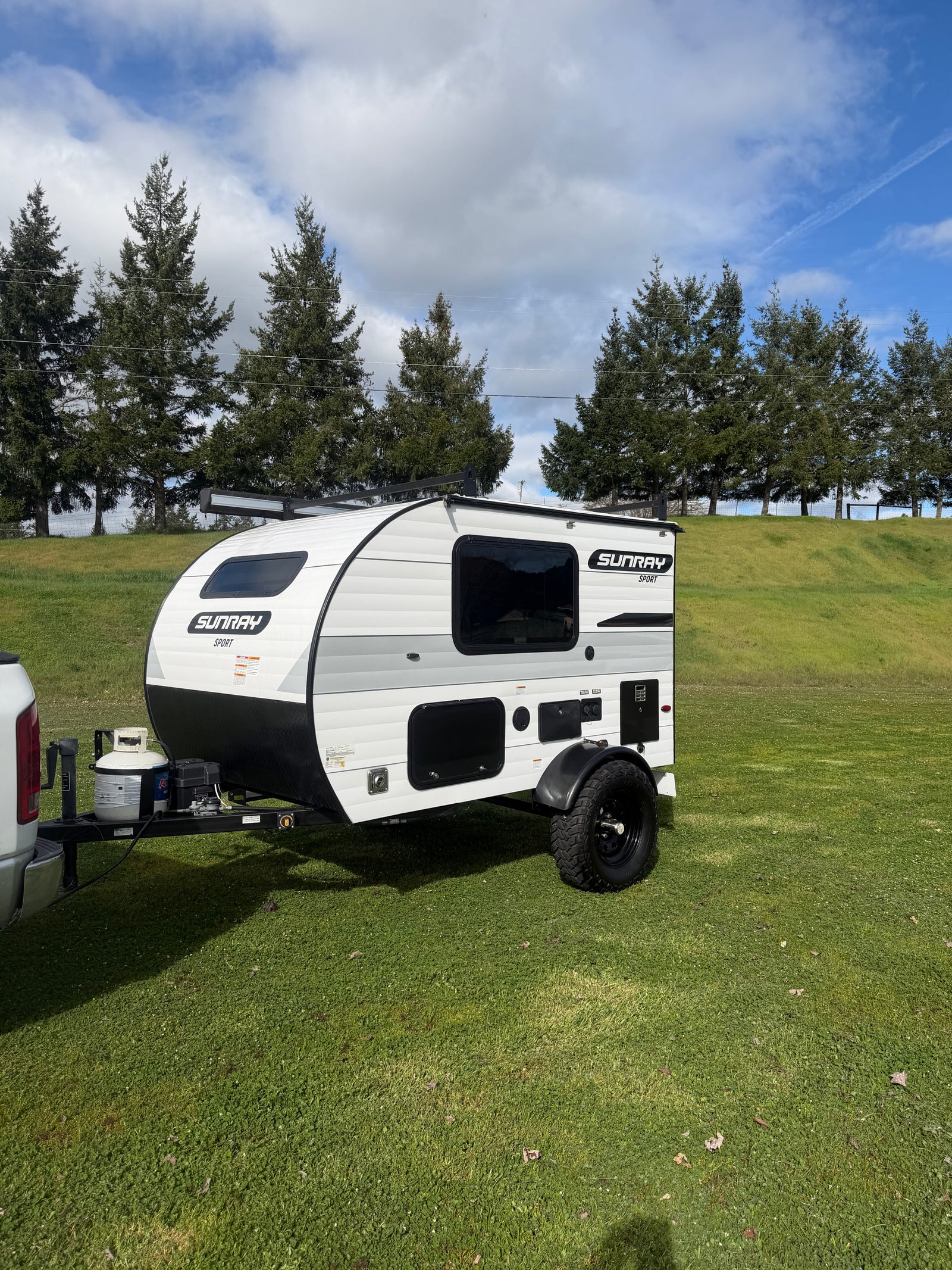 White Sunray Sport travel trailer for rv rental, parked on grass. Truck, trees, and cloudy sky in background.