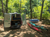 An RV rental camper van, doors open to reveal bed & storage, sits with kayaks by a lake, ready for campsite adventure.
