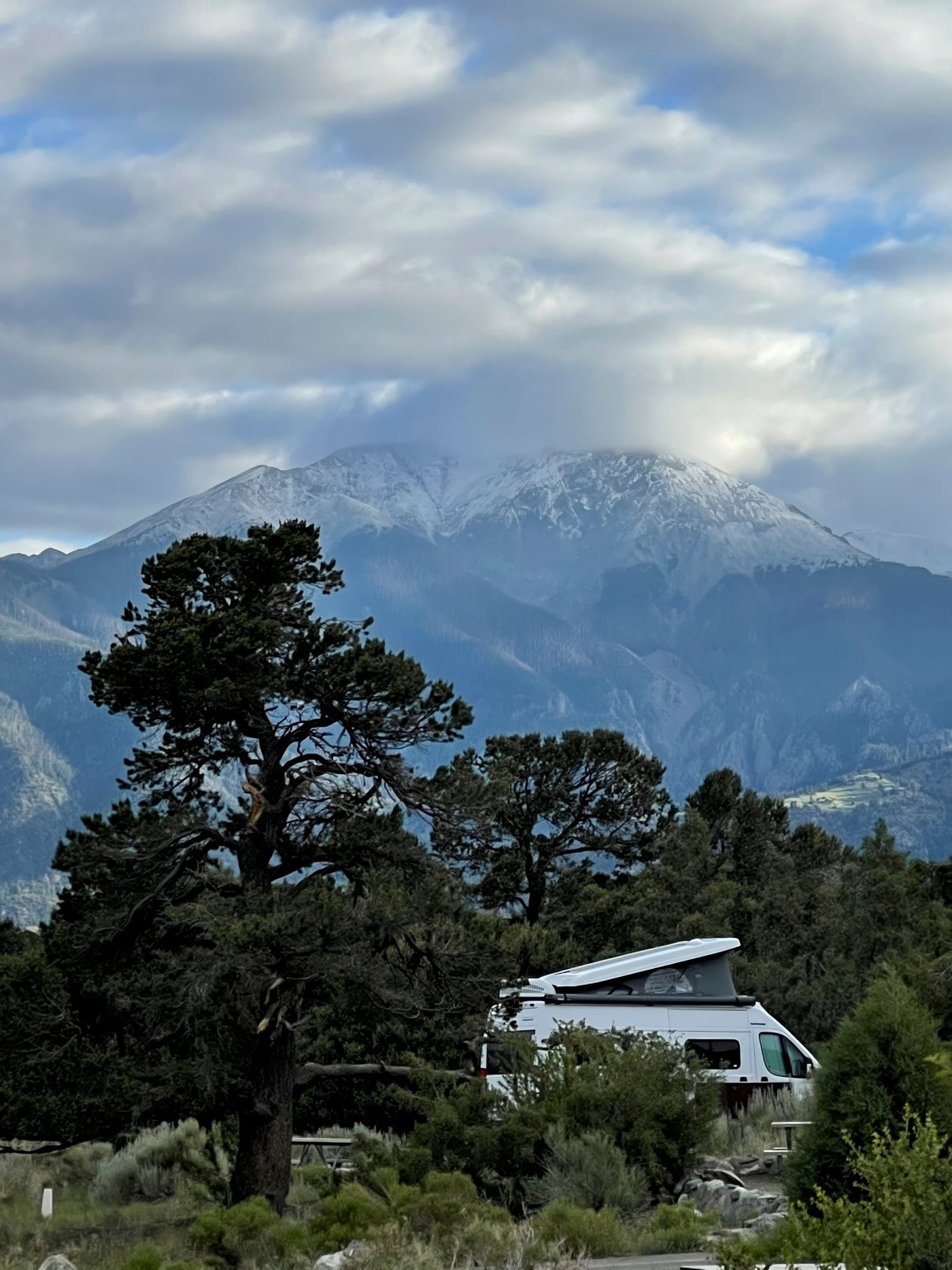 A white RV rental rests among green trees with a majestic, snow-capped mountain behind, under a cloudy sky. Ideal for nature adventures. — photo 5
