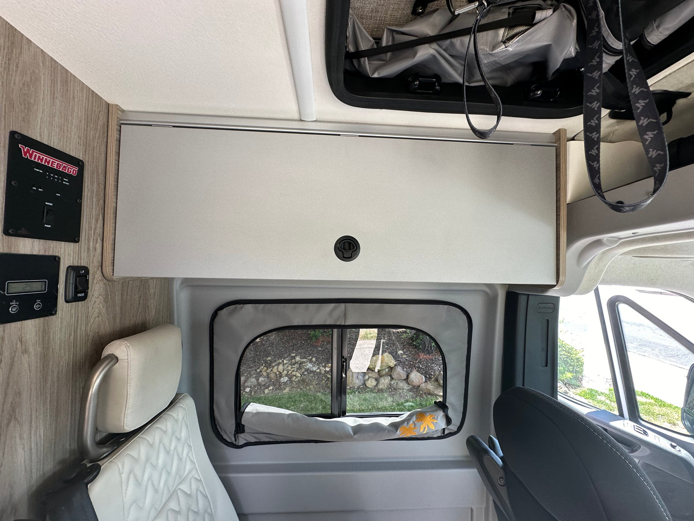 RV rental interior: A Winnebago camper van with light gray overhead storage, control panel, and a window shade featuring palm trees.
