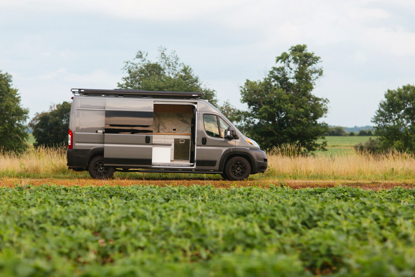 Custom grey camper van RV rental parked in fields, showing its open side door kitchen. Perfect for outdoor travel.