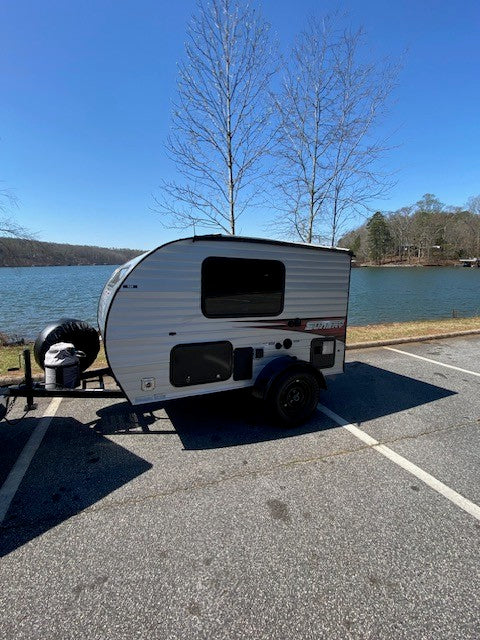 This compact RV rental, a small travel trailer, is parked by a scenic lake under a clear blue sky.