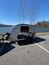 This compact RV rental, a small travel trailer, is parked by a scenic lake under a clear blue sky.