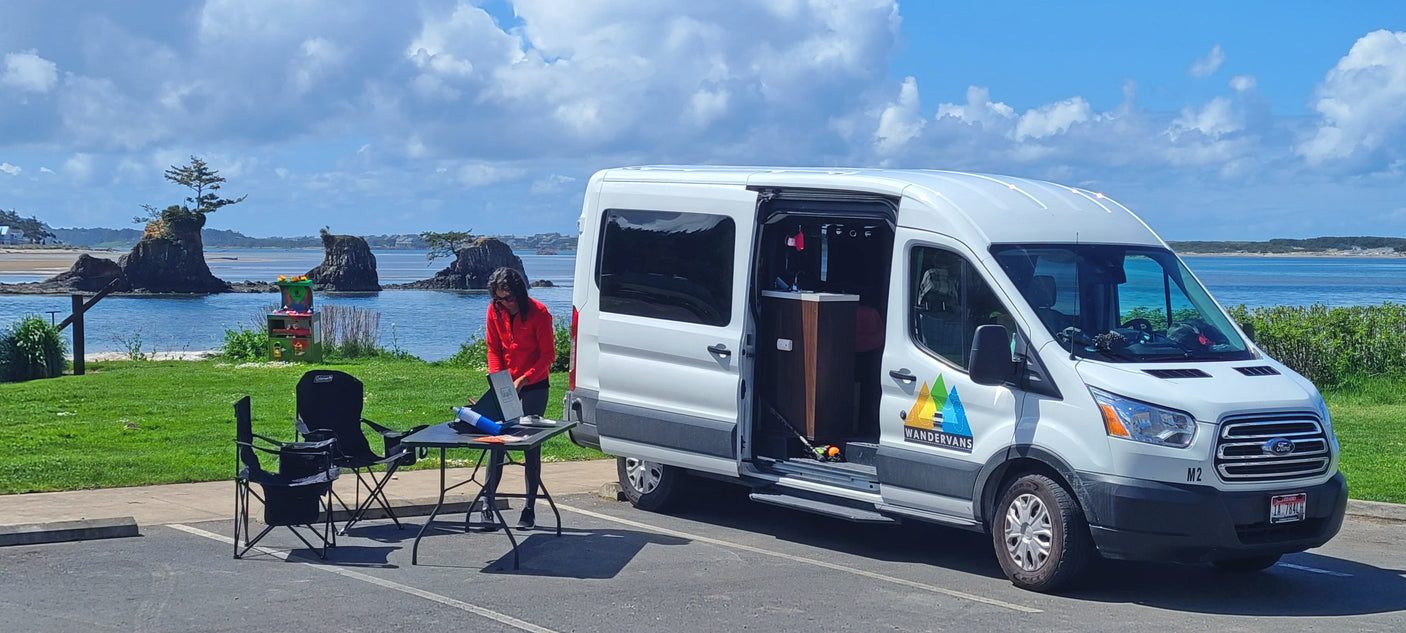 Wandervans RV rental campervan parked by a scenic coast. A person works on a laptop at a table, enjoying the outdoors.