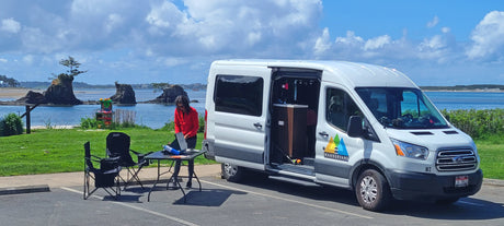 Wandervans RV rental campervan parked by a scenic coast. A person works on a laptop at a table, enjoying the outdoors.