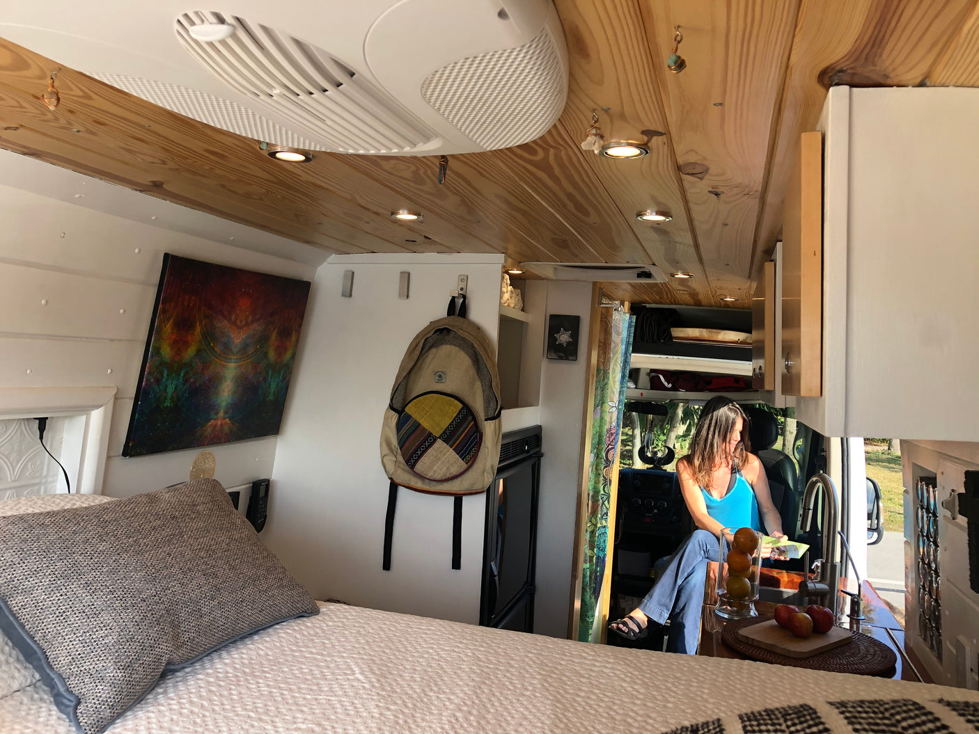 This RV rental's interior boasts a cozy bed, wood ceiling, and a woman relaxing near the compact kitchen and integrated cab area.