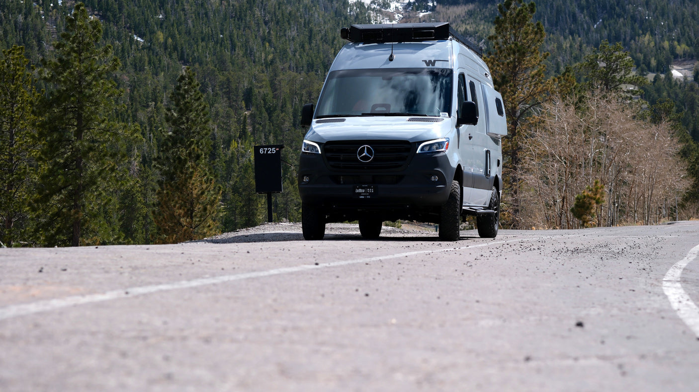An rv rental, a gray Mercedes-Benz camper van, drives on a paved mountain road through a pine forest.