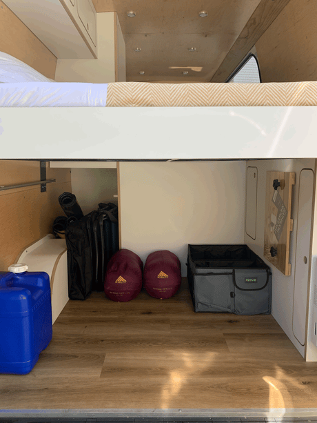 View into an RV rental's organized camper storage: sleeping bags, water jug, and gear neatly stowed under the bed.