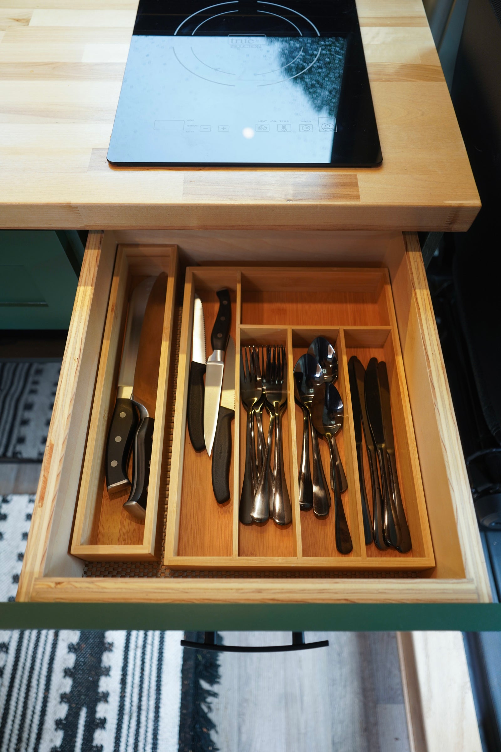 Top-down view of an RV rental kitchen: wood countertop, induction cooktop, and neatly organized cutlery drawer.