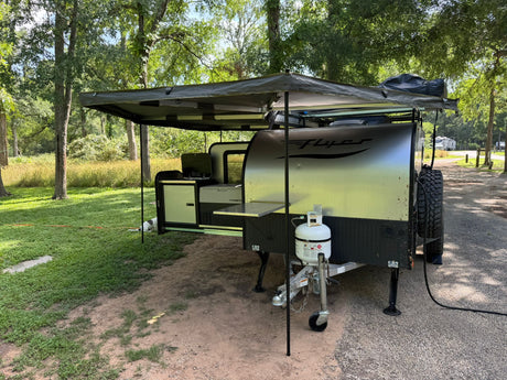 An RV rental, an off-road camper with an extended awning and outdoor kitchen, is set up at a leafy campsite.