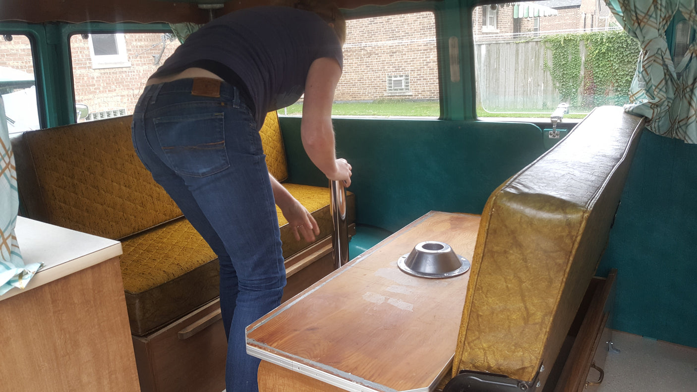 Inside an rv rental, a person adjusts a table base amid retro yellow seats and wood paneling.