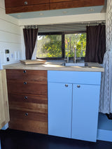 Custom RV rental kitchen featuring light wood counters, blue cabinets, sink, and a scenic window view.