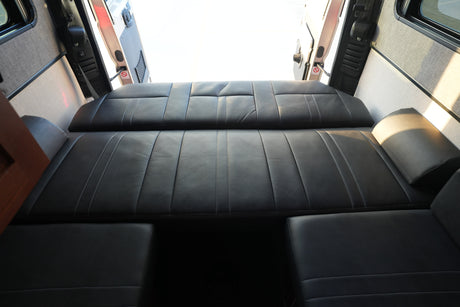 Spacious RV rental interior: black leather seating converts to a bed. Back doors open, bright light fills the camper van.