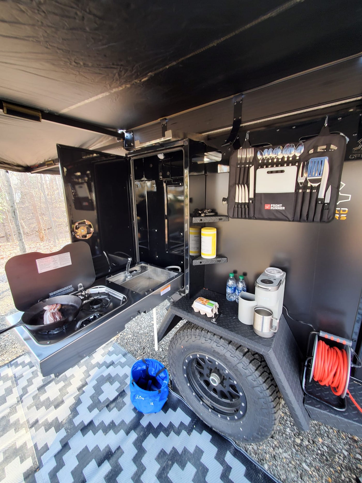  and a happy dog at a serene campsite.|A rugged black Expedition 2.0 off-road trailer with an open side revealing an integrated outdoor kitchen