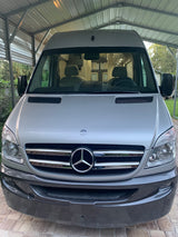 Silver Mercedes-Benz Sprinter camper van under a carport. Its front shows the large grille and headlights, ready for RV rental.