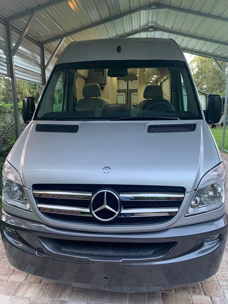 Silver Mercedes-Benz Sprinter camper van under a carport. Its front shows the large grille and headlights, ready for RV rental.