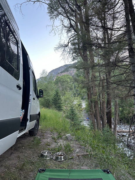 RV rental camper van parked by a mountain river, with pet bowls and a green cot on the ground, ideal for pet-friendly nature camping.