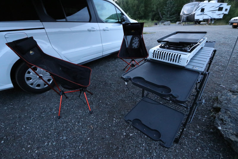 An RV rental van with a compact outdoor kitchen, griddle, and two black camping chairs on gravel. — photo 6