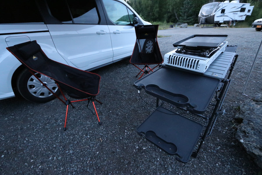 An RV rental van with a compact outdoor kitchen, griddle, and two black camping chairs on gravel.