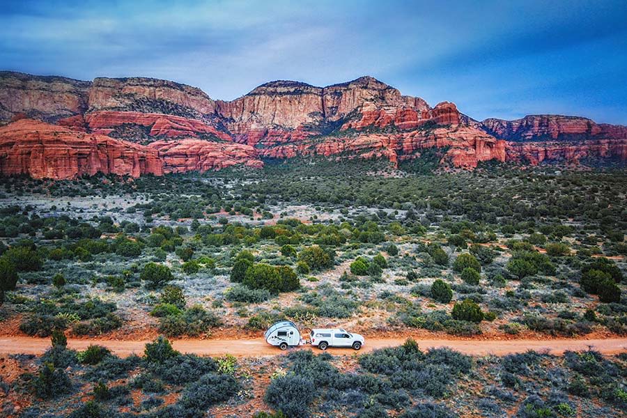 Scenic RV rental journey: a white SUV tows a teardrop camper on a dirt road, framed by stunning red rock mountains.