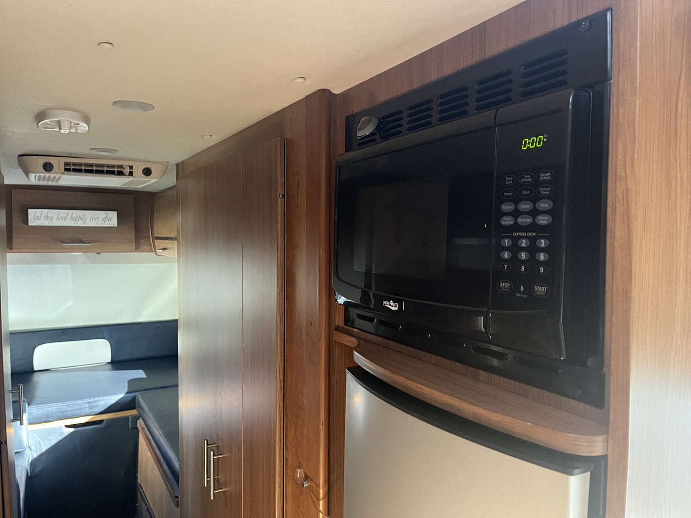 RV rental interior: Built-in microwave and fridge surrounded by warm wood cabinetry. Features an AC unit and cozy living space. — photo 20