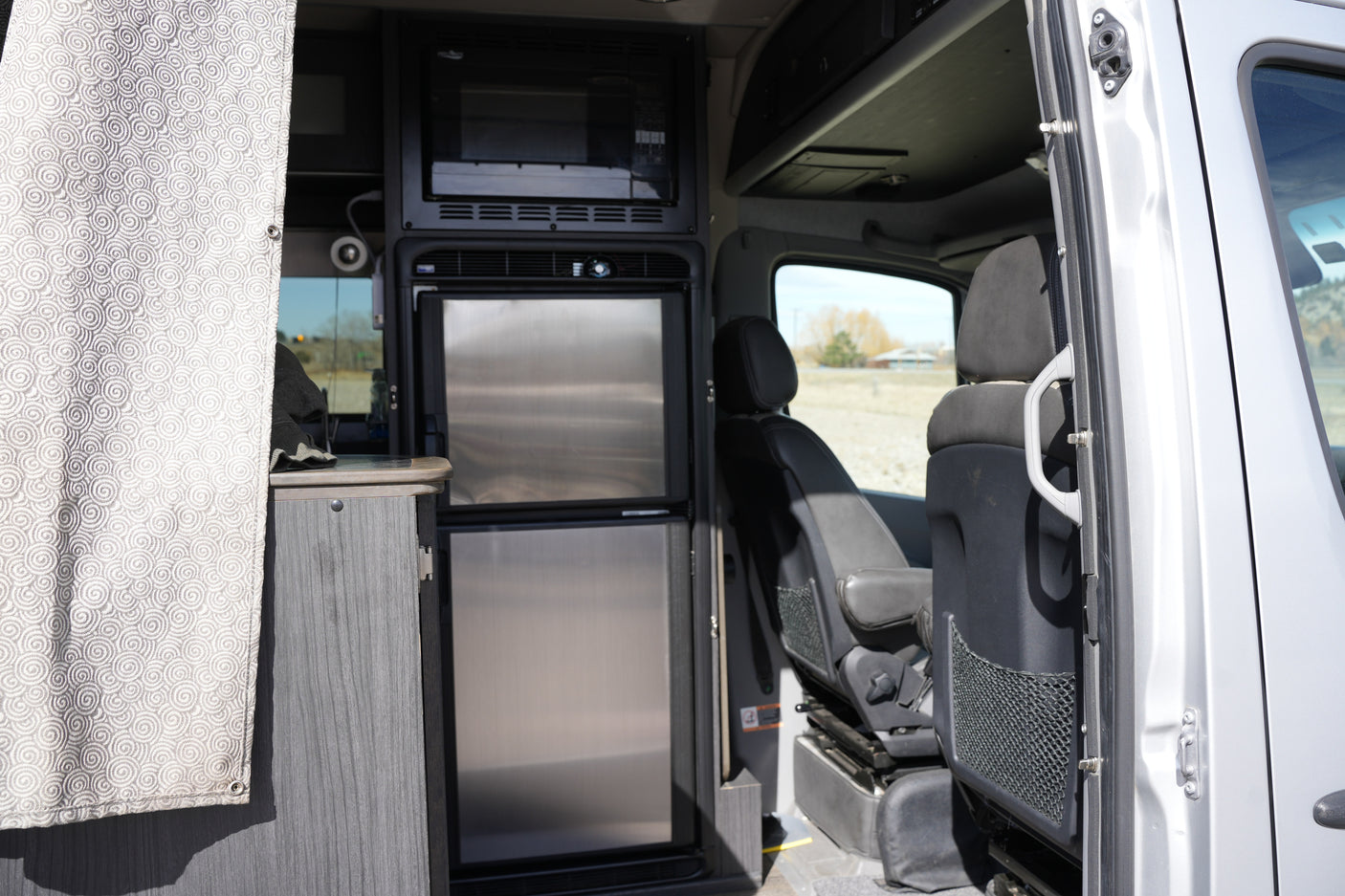 This RV rental interior displays a compact kitchen with microwave, two-door fridge, and the front cabin view through the open door.