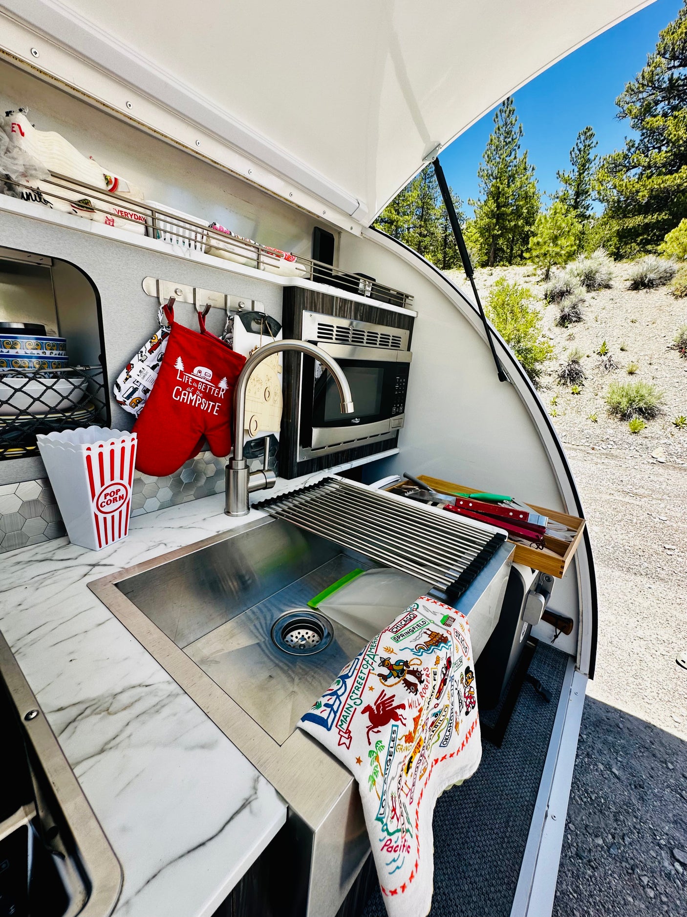 RV rental: compact outdoor kitchen with sink, microwave, "Life is Better at the Campsite" mitt, & nature views.